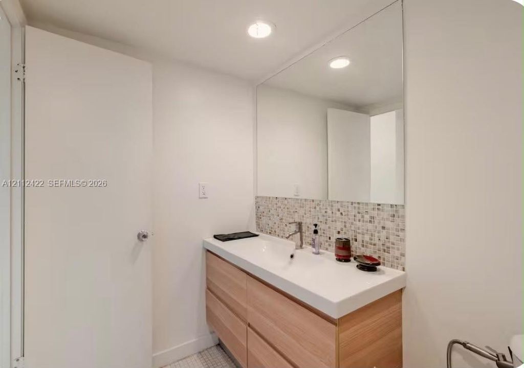 Floating wood-finish vanity with a rectangular sink, multi-toned mosaic tile backsplash, large wall-mounted mirror, and recessed ceiling lighting