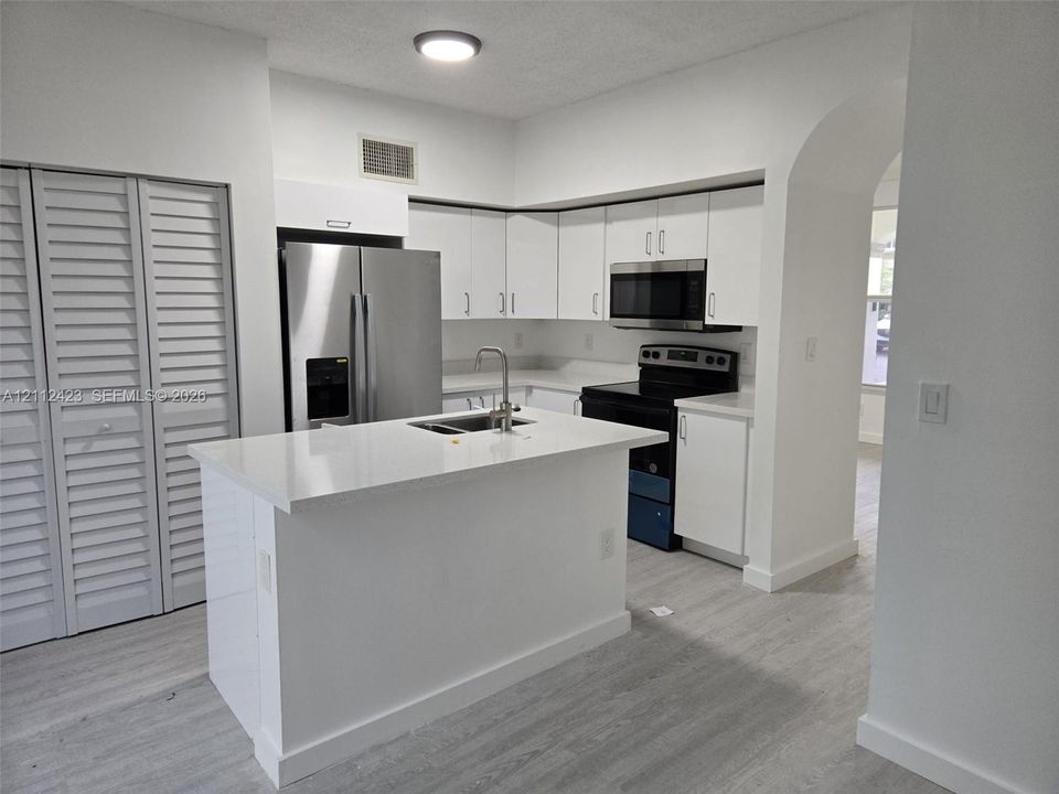 Kitchen featuring white shaker-style cabinetry, an island with an integrated stainless steel sink, stone-finish countertops, stainless steel refrigerator, and wood-finish flooring