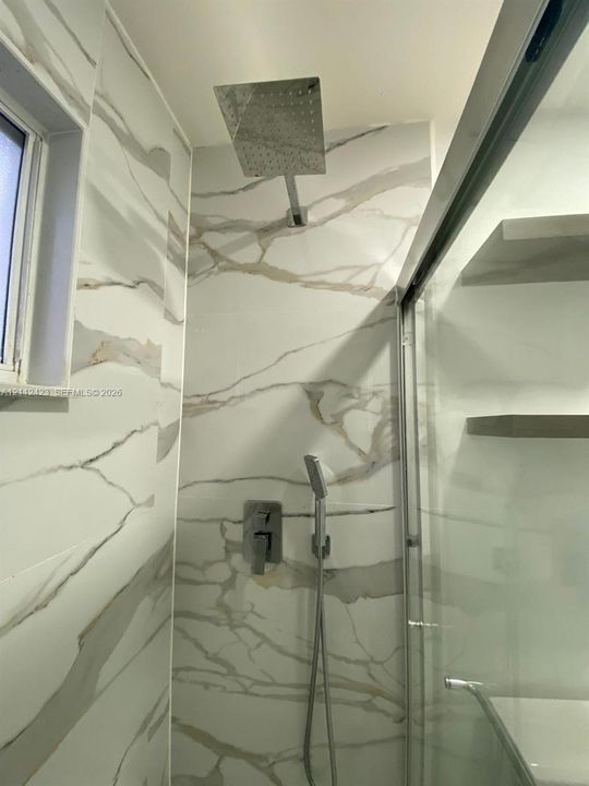 Shower enclosure featuring large-format veined tile surround, overhead rainfall showerhead, wall-mounted handheld sprayer, and built-in shelving