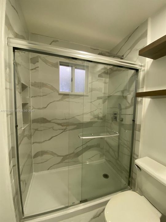 Walk-in shower enclosure featuring floor-to-ceiling veined tile surround, recessed wall niche, sliding glass doors, built-in shelving, and polished metal hardware