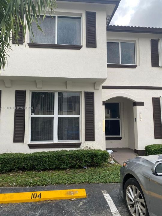 Two-story exterior featuring a light-toned stucco facade, dark decorative window shutters, an arched entryway, and manicured foundation shrubbery