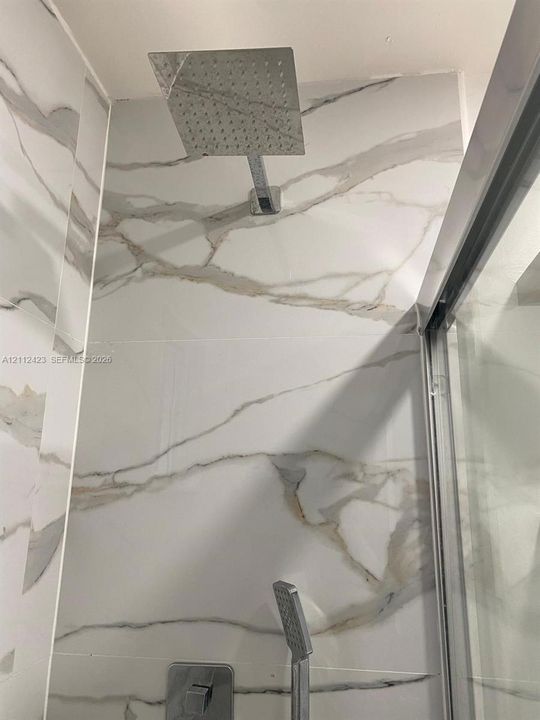 Walk-in shower featuring large-format wall tile with veining, a rainfall showerhead, a handheld shower wand, and a glass door enclosure