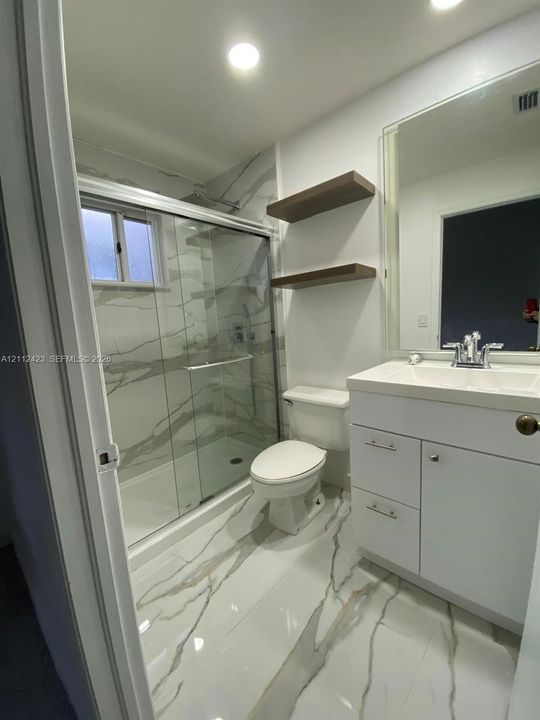Bathroom featuring a glass-enclosed shower with large-format veined tile surround, matching floor tile, white vanity with integrated basin, and two wood-finish floating shelves