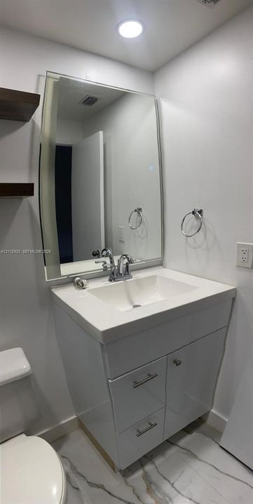 White vanity featuring an integrated rectangular sink, stone-finish countertop, polished chrome faucet, large rectangular mirror, and floating wall-mounted shelving