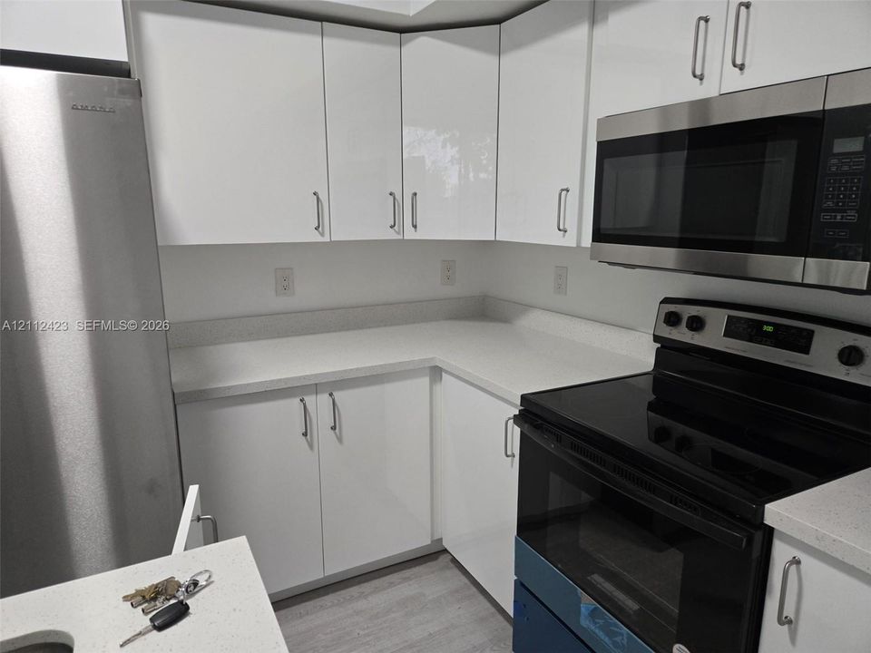 Gloss-finish white cabinetry, light-toned solid surface countertops, stainless steel refrigerator, stainless steel built-in microwave, and light-toned wood-finish flooring