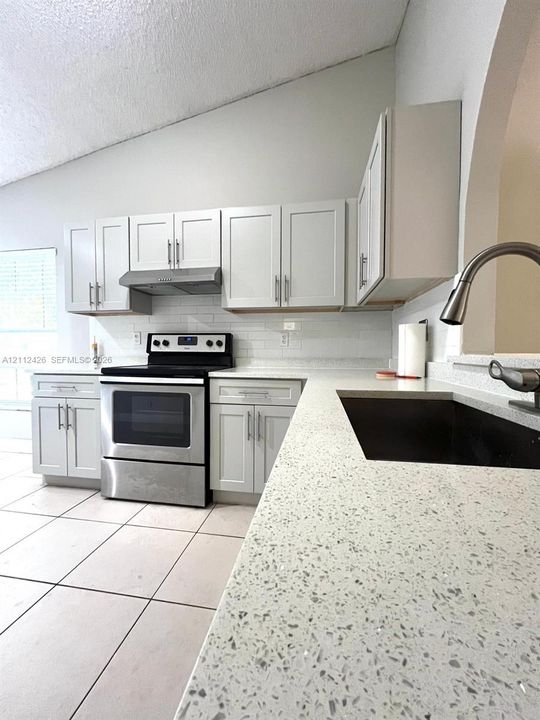 Kitchen featuring light-toned shaker cabinetry, stone-composite countertops, stainless steel appliances, a white tile backsplash, and ceramic floor tiling