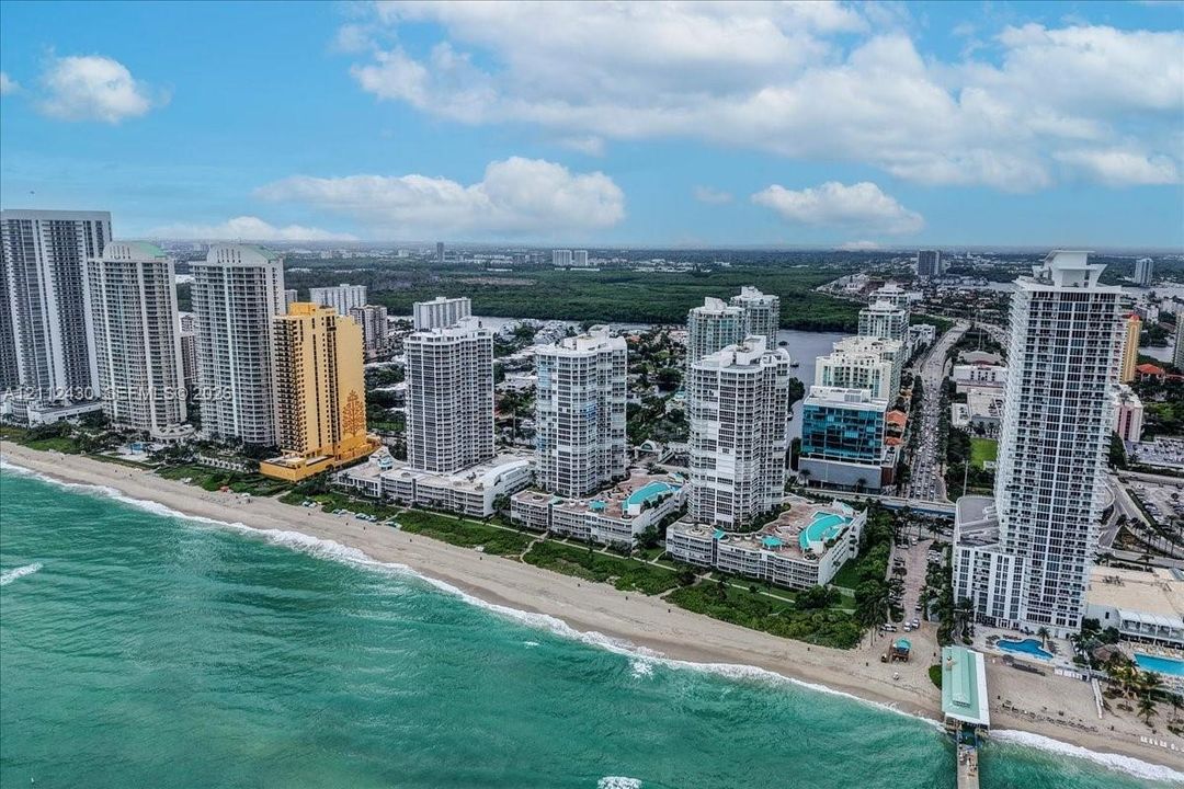 Aerial perspective of the coastline featuring oceanfront high-rise structures, sandy beach access, and tiered outdoor swimming pools