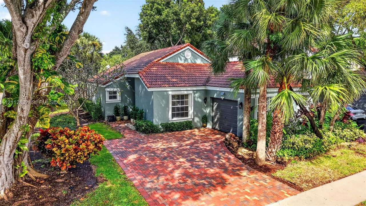 Single-story stucco exterior featuring a red tile roof, an expansive brick-paver driveway, and mature tropical landscaping