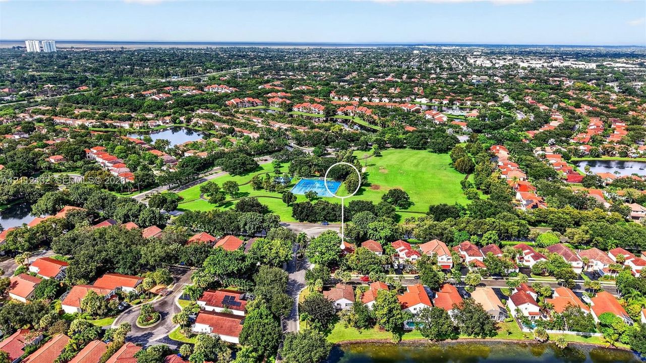 Aerial perspective featuring a residential property situated near an expansive green space, mature canopy trees, and community recreational court facilities