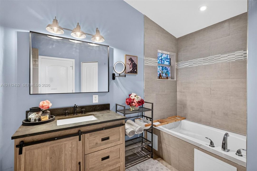 Wood-look vanity with a dark counter and rectangular drop-in sink, framed mirror with three-bulb vanity lighting, and a bathtub with tile surround and separate shower