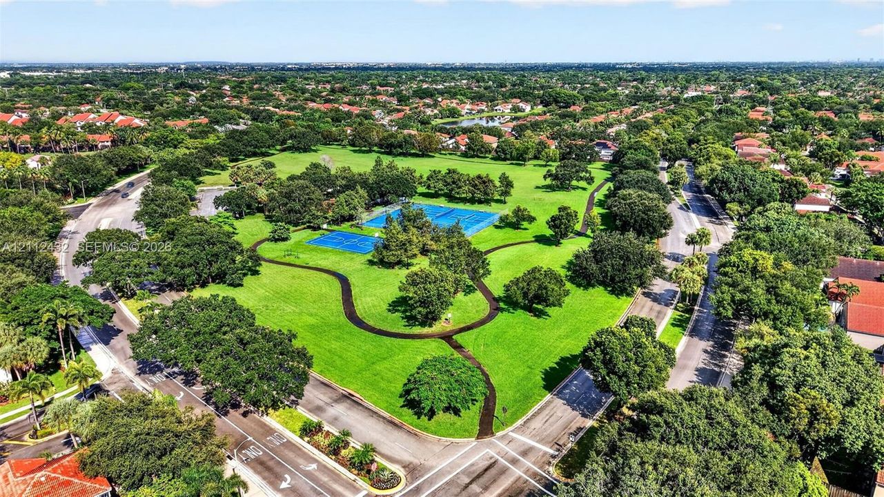 Aerial perspective of a sprawling park featuring multi-purpose paved recreation paths, blue-surface tennis courts, and mature landscaping