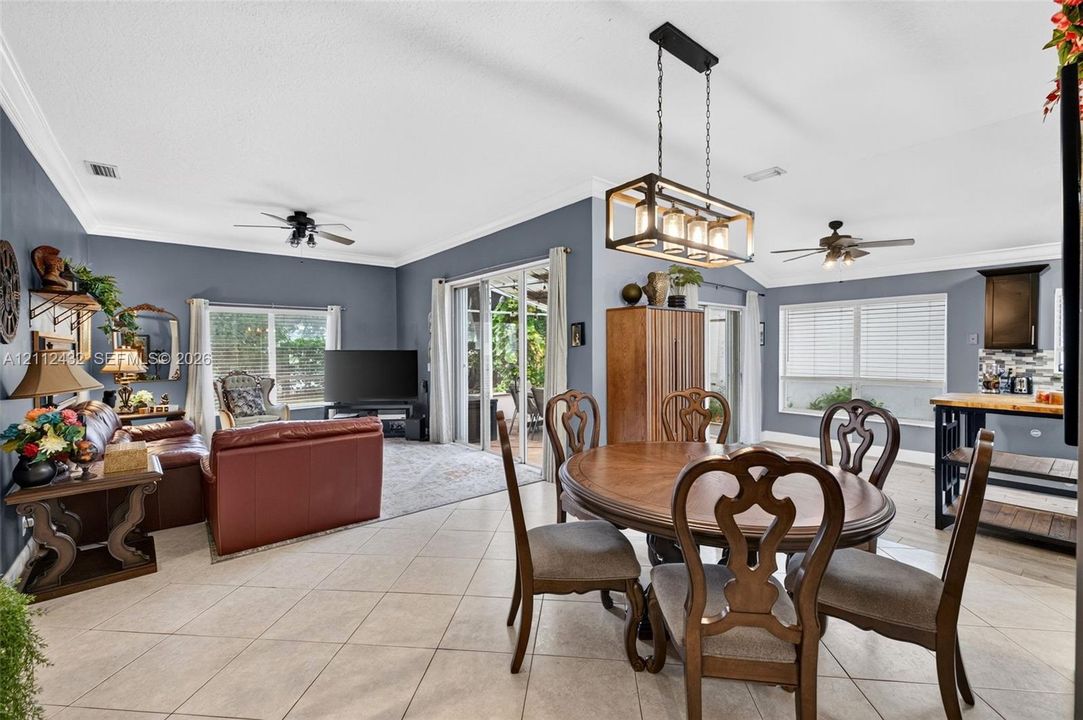 Open-concept main area featuring large-format ceramic tile flooring, crown molding, multi-light rectangular pendant fixture, dual ceiling fans, and multi-panel sliding glass doors