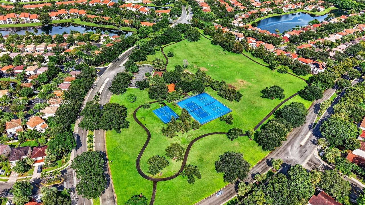 Expansive green space featuring tennis courts, a basketball court, mature trees, and paved walking paths