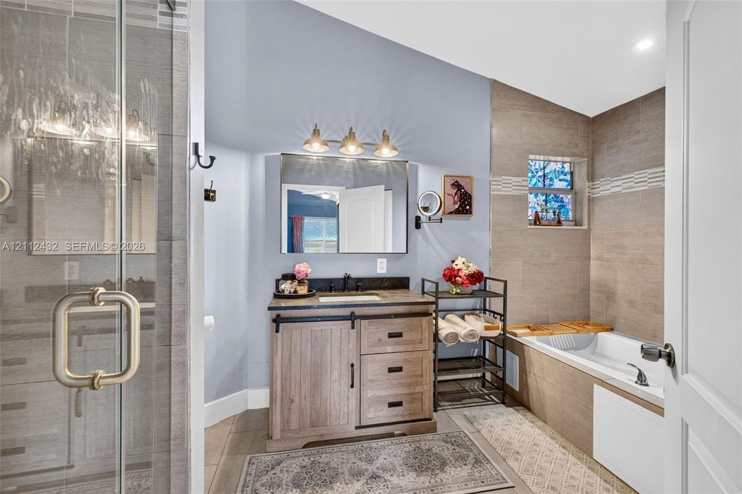 Bathroom featuring a wood-look vanity with a dark countertop, a glass-enclosed shower, a built-in soaking tub with tile surround, and a rectangular vanity mirror with integrated three-bulb lighting