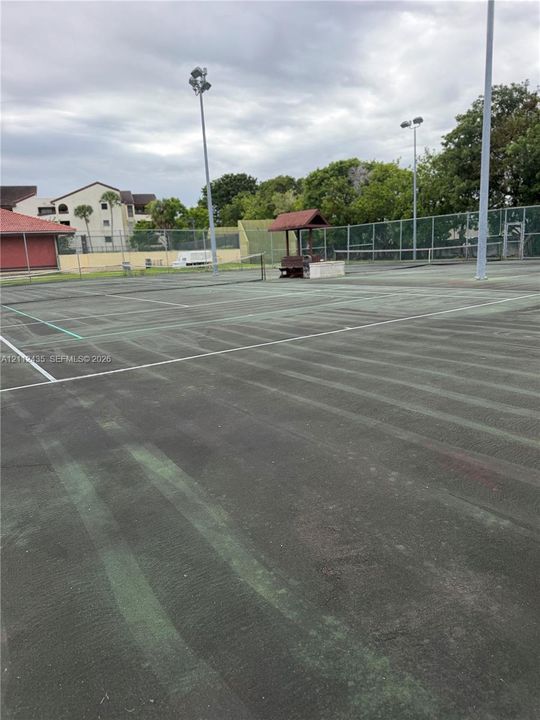 Tennis court with perimeter chain-link fencing, flood lighting, and a covered pavilion