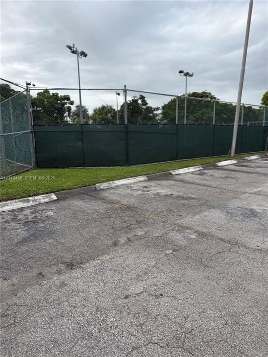 Asphalt parking area featuring concrete wheel stops, chain-link fencing, and overhead court lighting