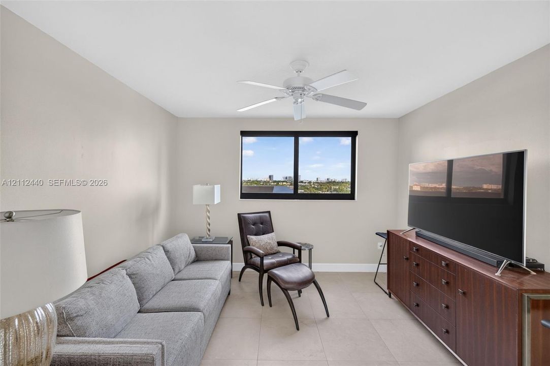 Neutral-toned guest bedroom featuring large-format porcelain flooring, a central ceiling fan with integrated lighting, and impact window with motorized roller shade