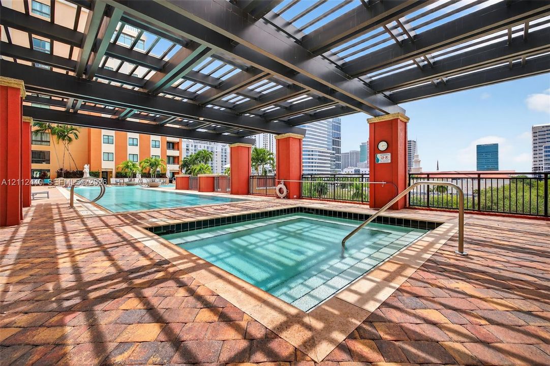 Covered pool and spa area featuring a dark-finish pergola, paver decking, and metal railings
