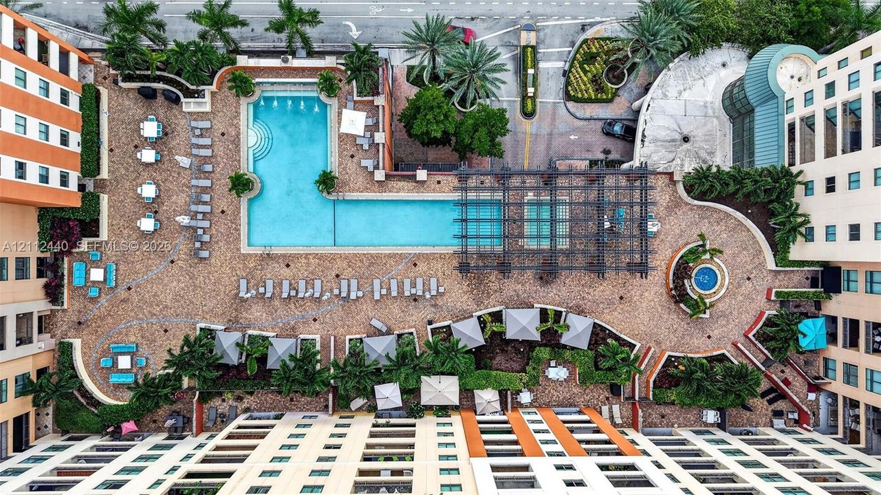 Aerial perspective of a sprawling outdoor terrace featuring an L-shaped swimming pool with fountains, a large pergola-covered area, patterned paver hardscaping, and professional landscaping with mature palm trees