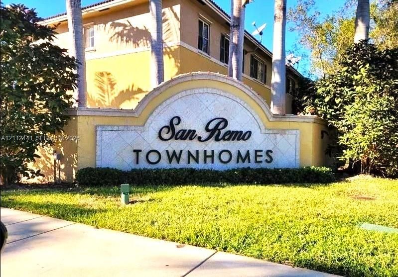San Remo Townhomes monument signage featuring a stucco arched wall, decorative tile inlay, and landscaped grounds with mature palms