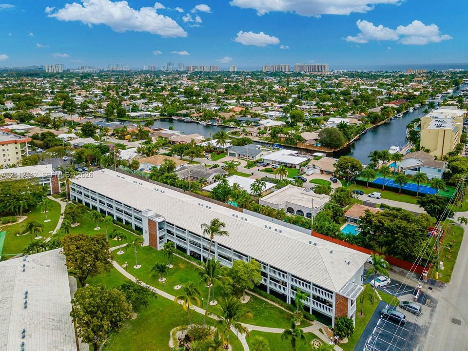 Aerial perspective featuring a multi-story residential building with a light-colored pitched roof, surrounded by landscaped grounds with mature palm trees, paved walkways, and dedicated surface parking areas