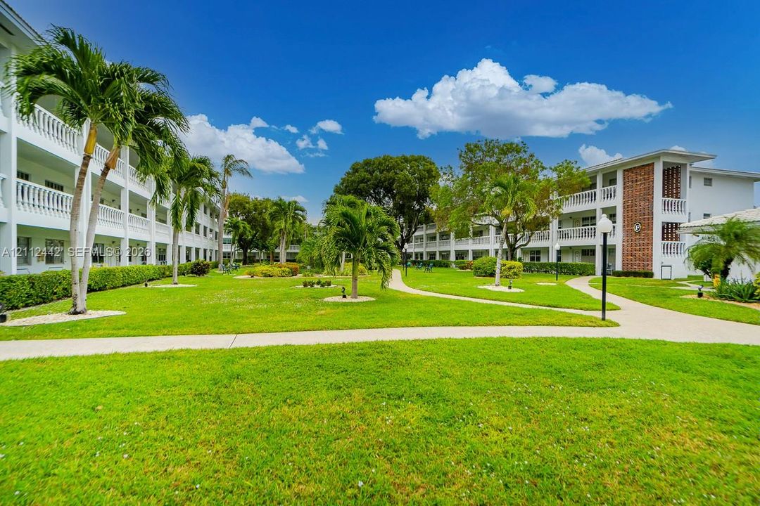 Expansive central courtyard featuring manicured lawn, paved walkways, mature palm trees, and multi-story buildings with white balcony railings
