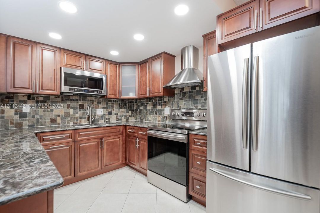 Kitchen featuring cherry-tone cabinetry, stone-patterned countertops, mosaic tile backsplash, stainless steel appliances, and a professional-grade range hood