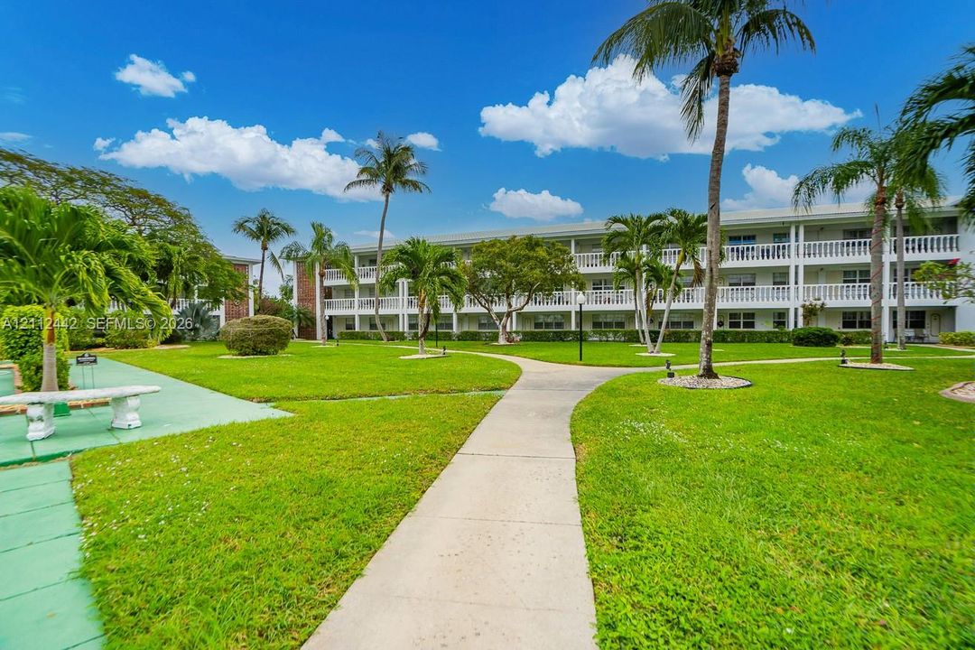 Mid-rise multi-story building exterior featuring open-air corridors with decorative balustrade railings, manicured green lawns, paved walkways, and mature palm trees