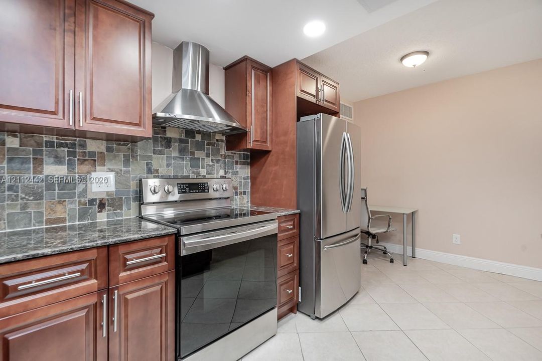 Kitchen featuring stained wood cabinetry, stone-tiled backsplash, polished stone countertops, stainless steel appliances, and tile flooring