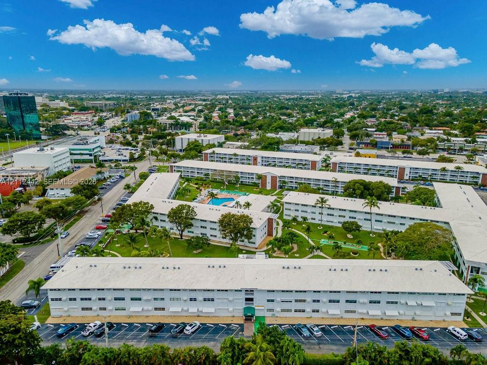 Aerial perspective of a multi-building residential complex featuring white exterior facades, an in-ground swimming pool, manicured grounds, and dedicated surface parking