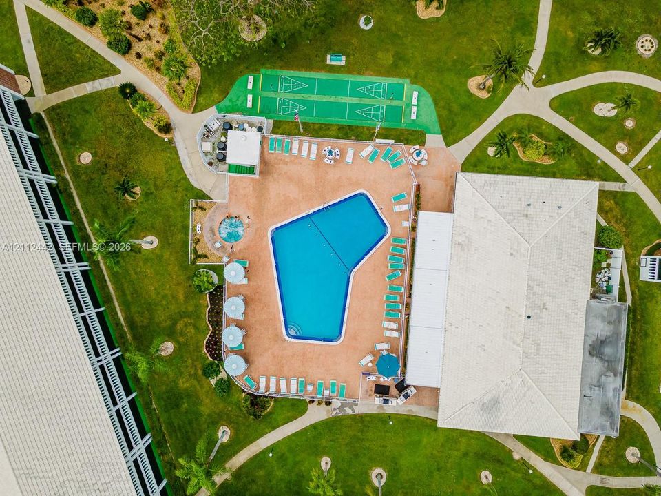 Aerial perspective of a common area featuring a swimming pool, integrated spa, paved sun deck, and shuffleboard courts set within landscaped grounds