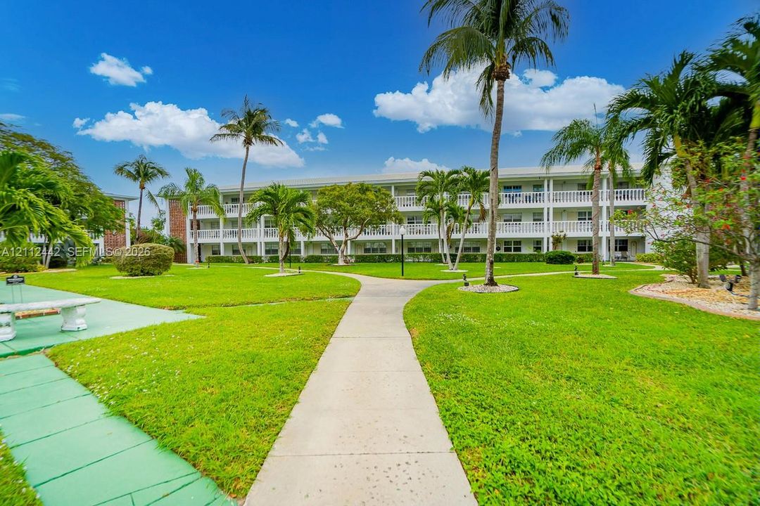 Expansive green space featuring mature palm trees, manicured landscaping, concrete pathways, and a multi-story building with exterior balconies