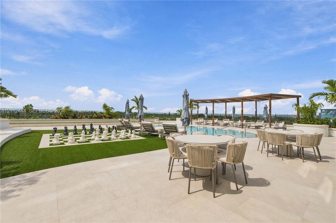 Outdoor terrace featuring light-toned stone paving, a swimming pool, a wood-frame pergola, synthetic turf, and an oversized permanent outdoor chess board