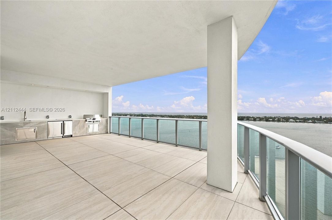 Covered terrace featuring large-format tile flooring, a built-in outdoor kitchen with stone-finish cabinetry, an integrated grill, and a glass-paneled railing