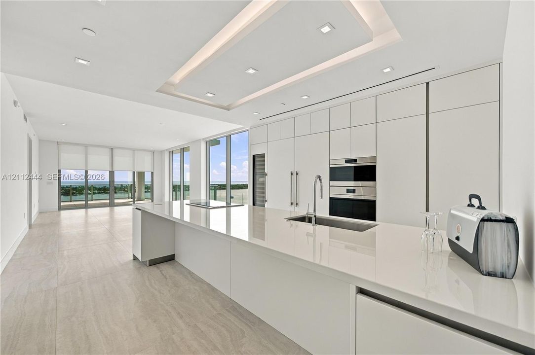Modern kitchen featuring seamless flat-panel cabinetry, a spacious solid-surface island, built-in stainless steel appliances, recessed tray ceiling lighting, and large-format tile flooring