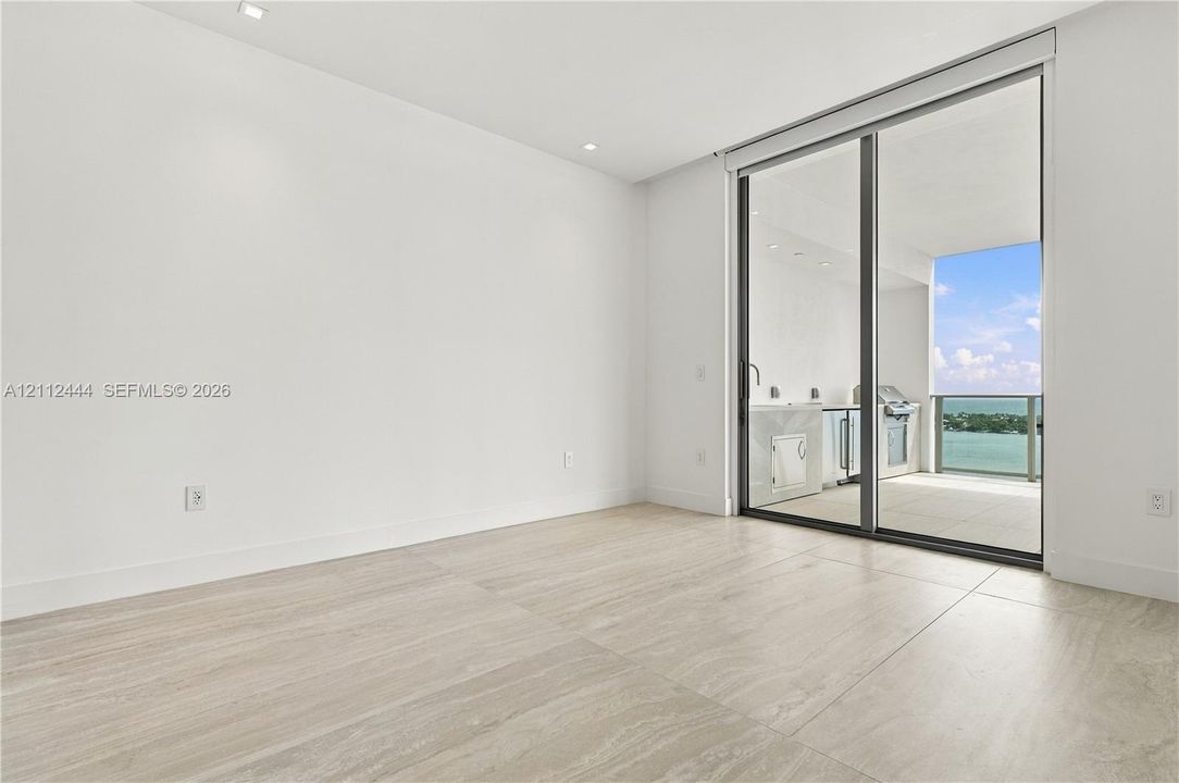 Large-format tile flooring, recessed ceiling lighting, and floor-to-ceiling sliding glass doors opening to a covered balcony with built-in cabinetry