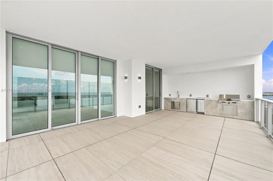 Expansive covered terrace featuring large-format tile flooring, a built-in outdoor kitchen with stainless steel appliances, stone-finish cabinetry, floor-to-ceiling glass sliding doors, and recessed lighting