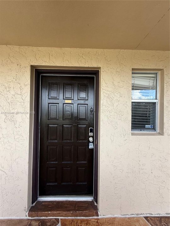 Dark-finished multi-panel entry door with electronic deadbolt, textured stucco exterior, and window with integrated blinds