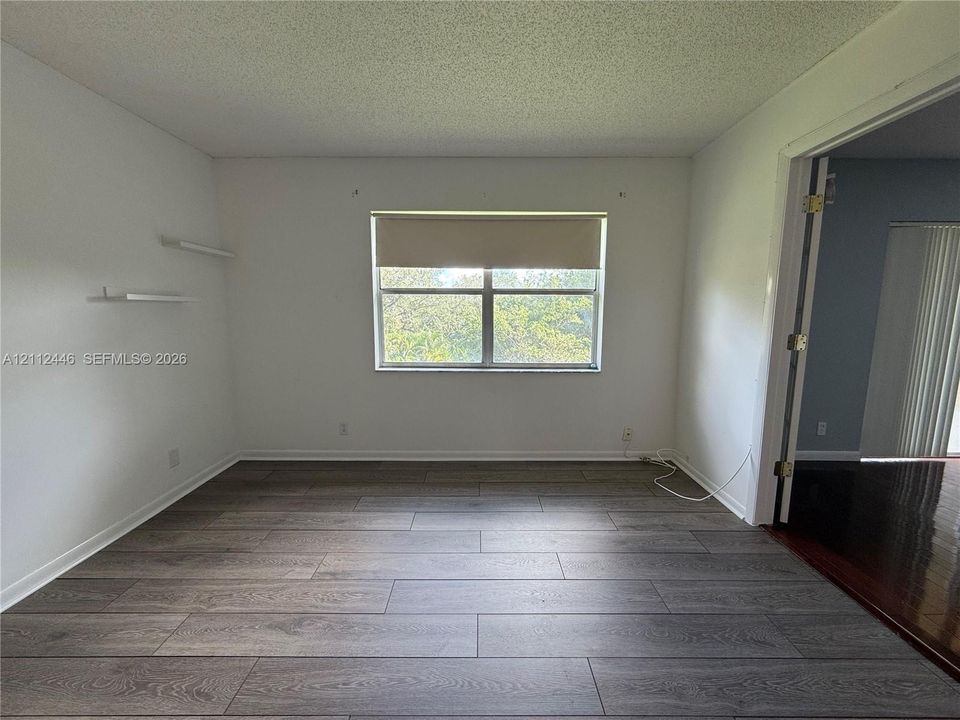 Interior room featuring wood-finish flooring, two wall-mounted shelves, and a single-pane window with a roller shade