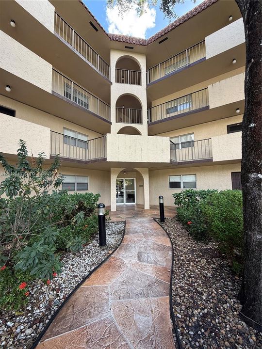 Multi-story stucco exterior featuring arched architectural detailing, private balconies with metal railings, a tiled roofline, and a flagstone walkway