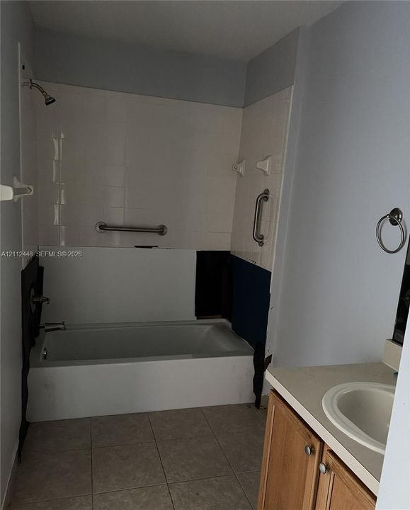 Bathtub with wall-mounted shower head, tile surround, and installed safety grab bars, paired with a wood-finish vanity featuring an integrated basin and a neutral countertop