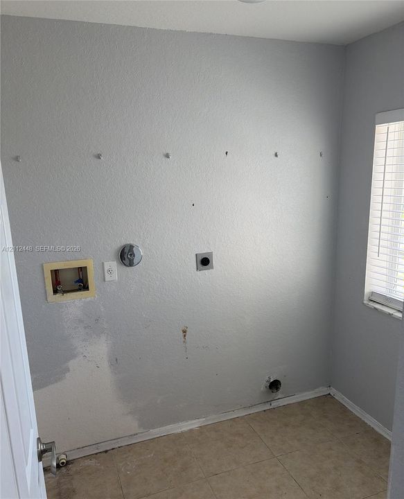 Dedicated utility space featuring tile flooring, a window with horizontal blinds, and existing plumbing and electrical hookups