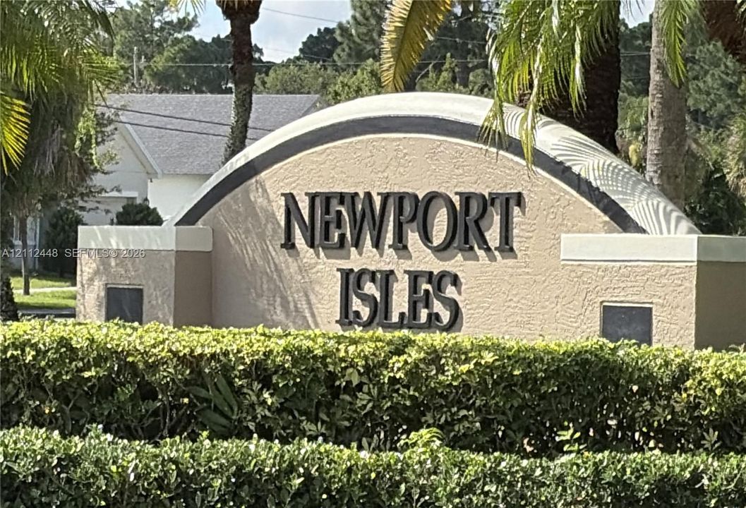 Community monument sign featuring stucco construction, arched detailing, and elevated lettering, accented by mature perimeter hedging and palm trees