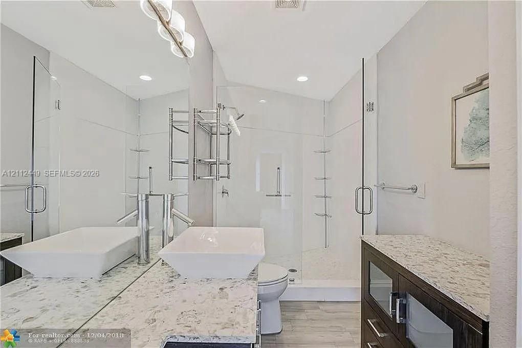 Bathroom featuring a dual-sink vanity with light-toned stone countertops, a frameless glass-enclosed shower, wood-finish flooring, and a full-width wall mirror