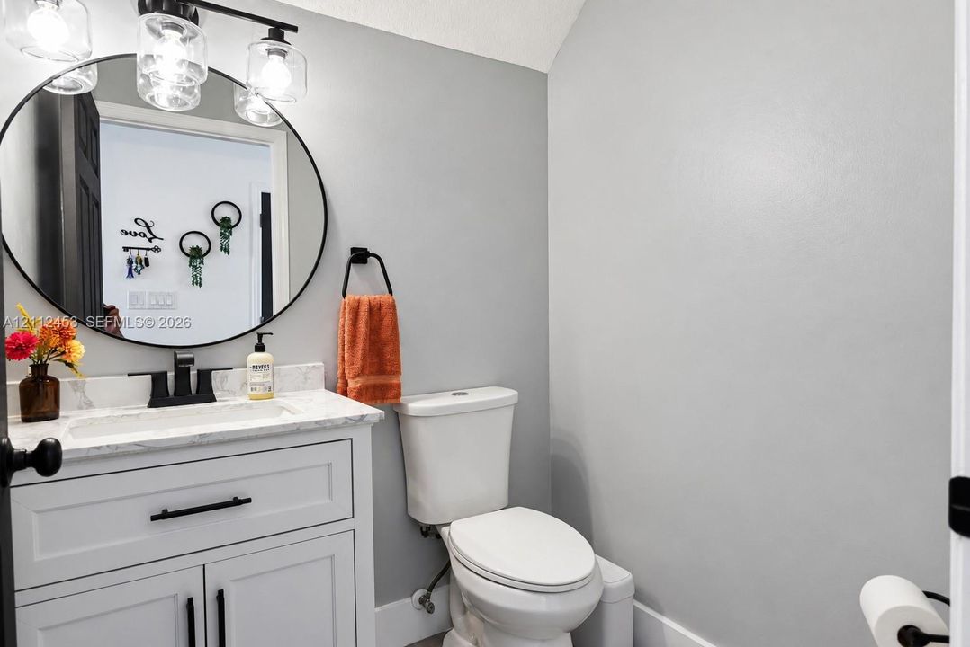 White vanity with a stone-composite countertop, integrated rectangular sink, matte black hardware, circular mirror, and multi-light vanity fixture