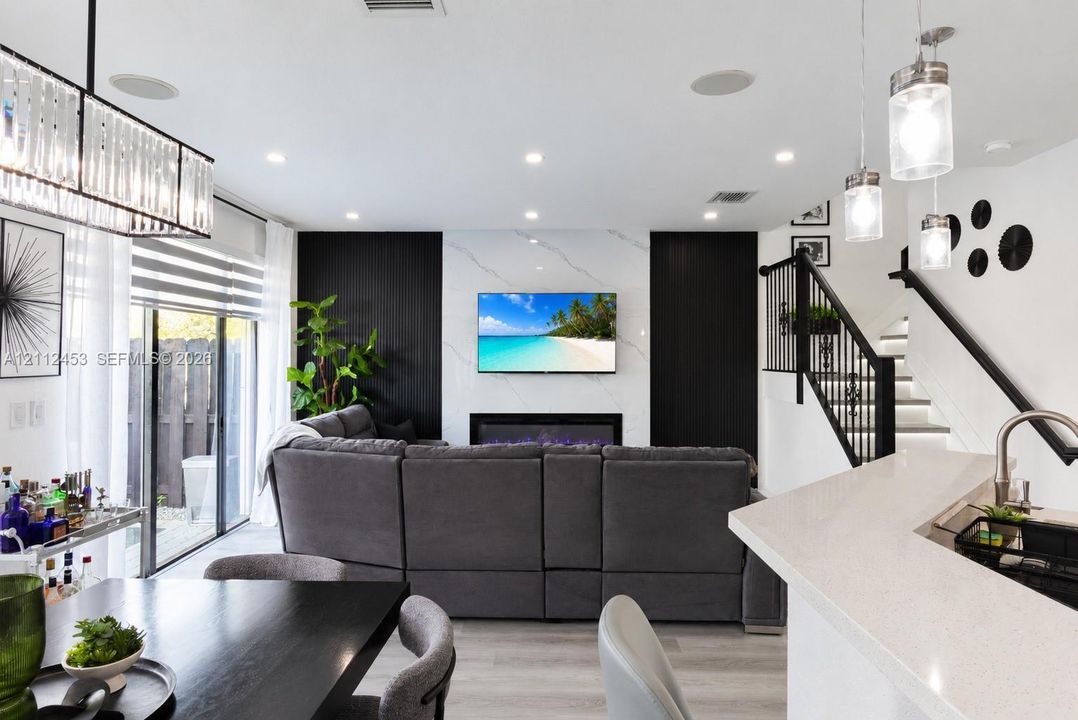 Living area featuring a floor-to-ceiling fireplace surround with stone-finish cladding, recessed ceiling lighting, a modern chandelier, and a staircase with matte black railings