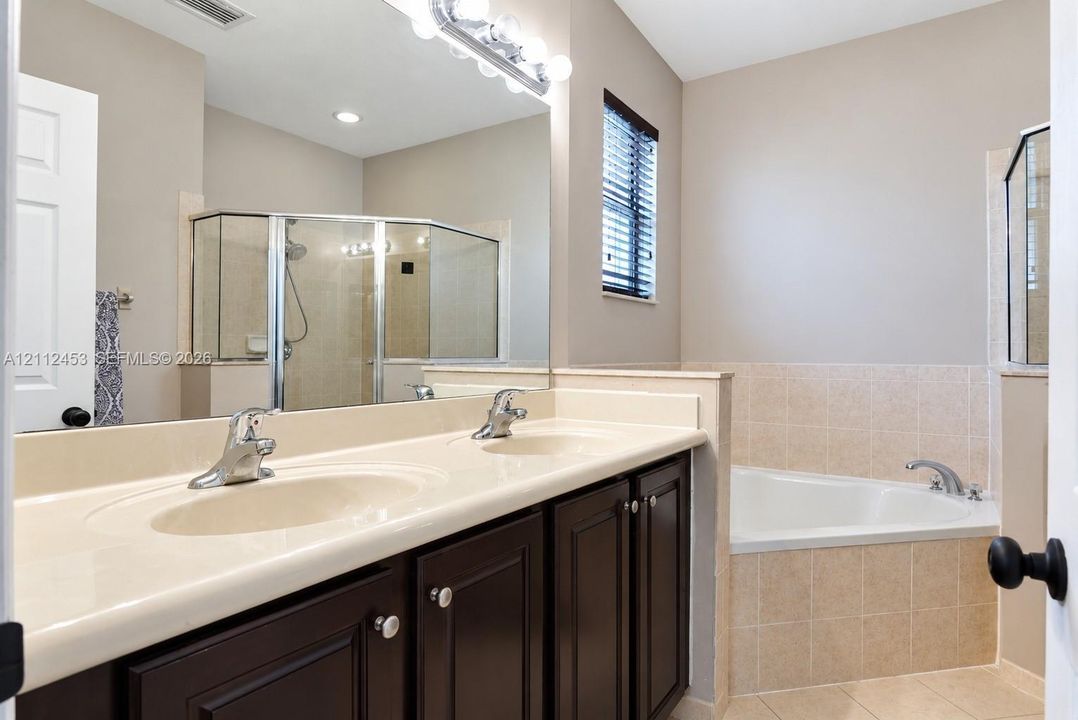 Double-sink vanity with a solid-surface countertop, dark-finish cabinetry, corner soaking tub with tiled surround, and separate glass-enclosed shower