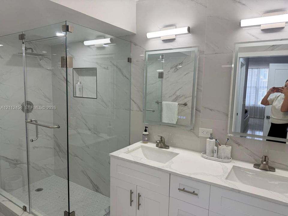 Dual-sink vanity with white cabinetry and stone countertop, glass-enclosed walk-in shower with integrated wall niche, floor-to-ceiling stone-style wall tile, and wall-mounted light fixtures