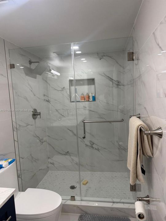 Walk-in shower featuring a frameless glass enclosure, floor-to-ceiling stone-pattern tile, integrated recessed shelving, polished metal hardware, and a pebble-style basin floor