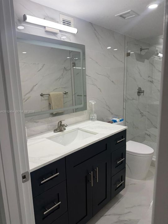 Dark-finish vanity with a rectangular sink, stone-surface countertop, LED-backlit mirror, and full-height stone-patterned wall tile